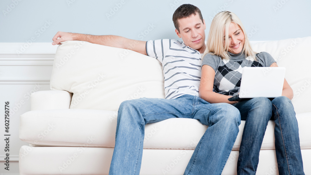 Man and woman sit side by side on a white couch. The woman is using a laptop, and the man is snuggling next to her. Horizontal format. Man and woman sit side by side on a white couch. The woman is using a laptop, and the man is snuggling next to her. Horizontal format.