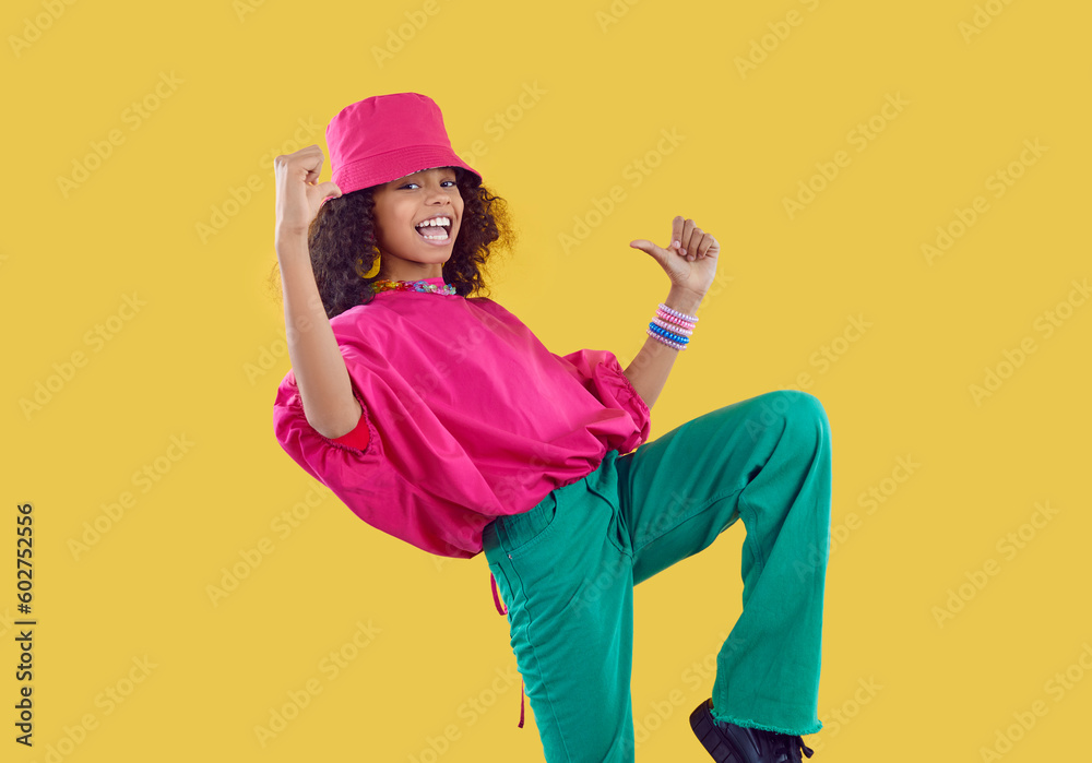 Little kid in trendy clothes. Happy child having fun in fashion studio. Afro American girl wearing fuschia bucket hat, loose oversized top and green jeans dancing isolated on solid yellow background - obrazy, fototapety, plakaty 