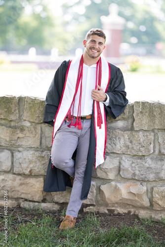 Smiling, confident University of Louisville college graduate wearing a gown and honors ropes leaning on stone retaining wall in the park photo