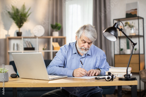 Confident caucasian man of mature age taking notes while utilizing portable computer in remote work from home. Skillful freelancer in business casual clothes writing initial report of project indoors. © sofiko14