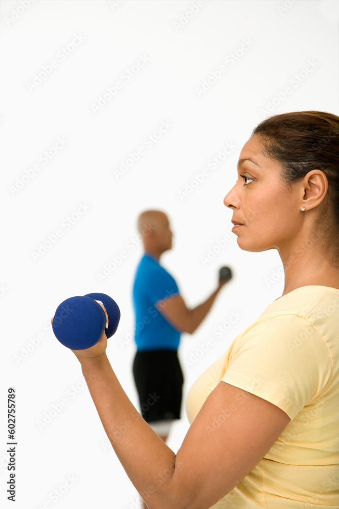 Close up of smiling mid adult multiethnic woman exercising using dumbbells with mid adult multiethnic man in background. Close up of smiling mid adult multiethnic woman exercising using dumbbells with mid adult multiethnic man in background.