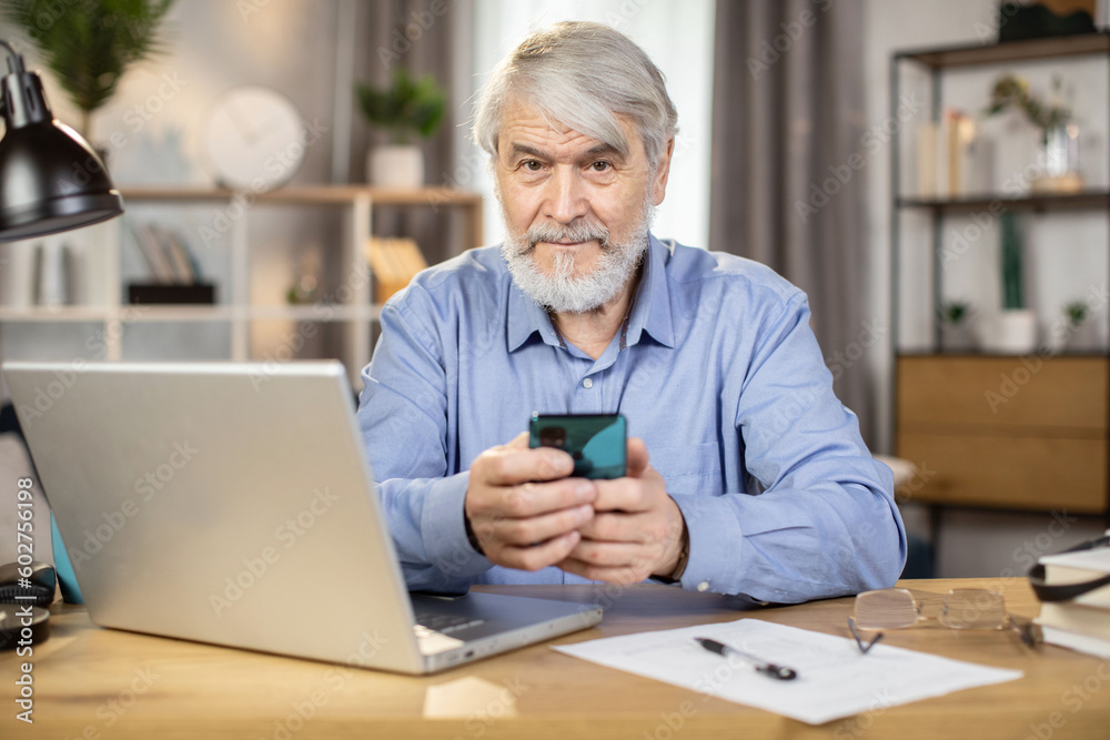 Mindful caucasian man of mature age holding cell phone while teleworking in home interior. Professional business leader looking through contact information on smart gadget at desk in remote office. Mindful caucasian man of mature age holding cell phone while teleworking in home interior. Professional business leader looking through contact information on smart gadget at desk in remote office.