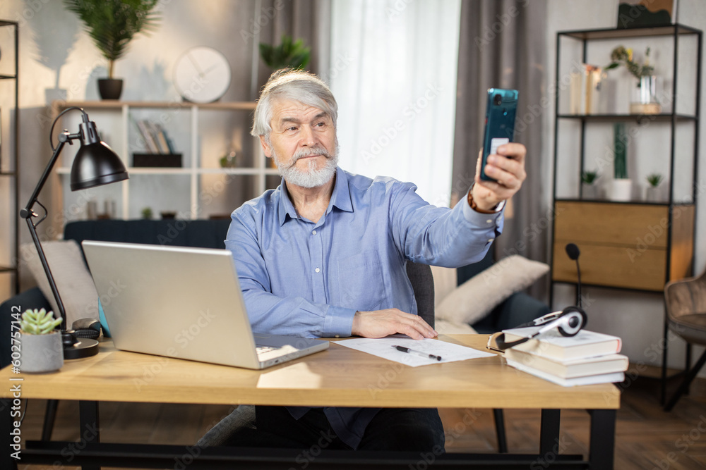 Smiling man about 65 posing with pointing finger in front of smartphone camera while taking break from work in home office. Mature freelancer taking selfie of comfortable lifestyle image indoors. Smiling man about 65 posing with pointing finger in front of smartphone camera while taking break from work in home office. Mature freelancer taking selfie of comfortable lifestyle image indoors.