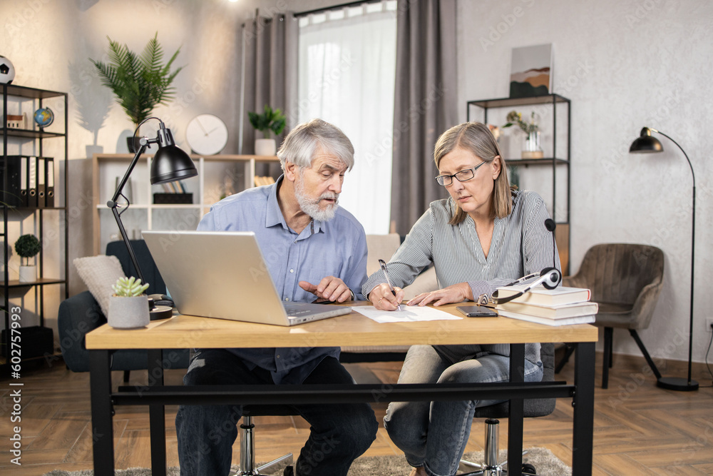 Middle-aged lady in glasses putting down information while senior male finding data on laptop in creative office. Efficient professionals drafting written agreement while doing distant work from home. Middle-aged lady in glasses putting down information while senior male finding data on laptop in creative office. Efficient professionals drafting written agreement while doing distant work from home.