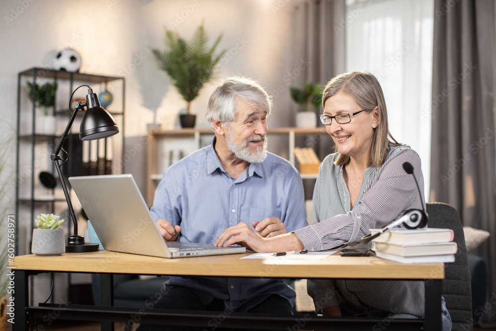 Elegant senior lady in glasses and gray-haired elderly man utilizing portable computer for distant work at home. Efficient freelancers studying details of temporary contract on digital screen.