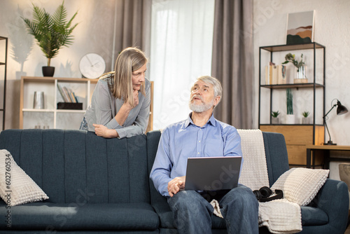 Принти на полотні Beautiful mature lady helping senior husband with video call app on laptop while standing behind couch in living room