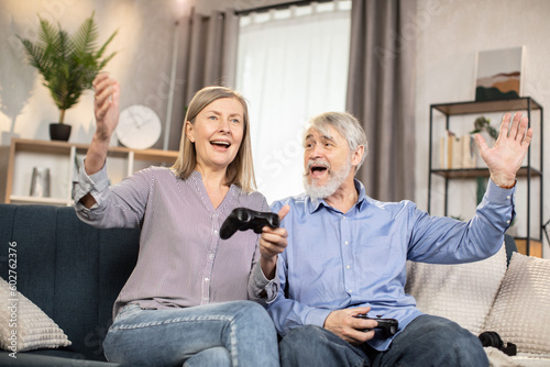 Excited man and woman of mature age holding black gamepads while playing on console at home. Emotional married couple in casual clothes expressing joy about successful result of video entertainment.