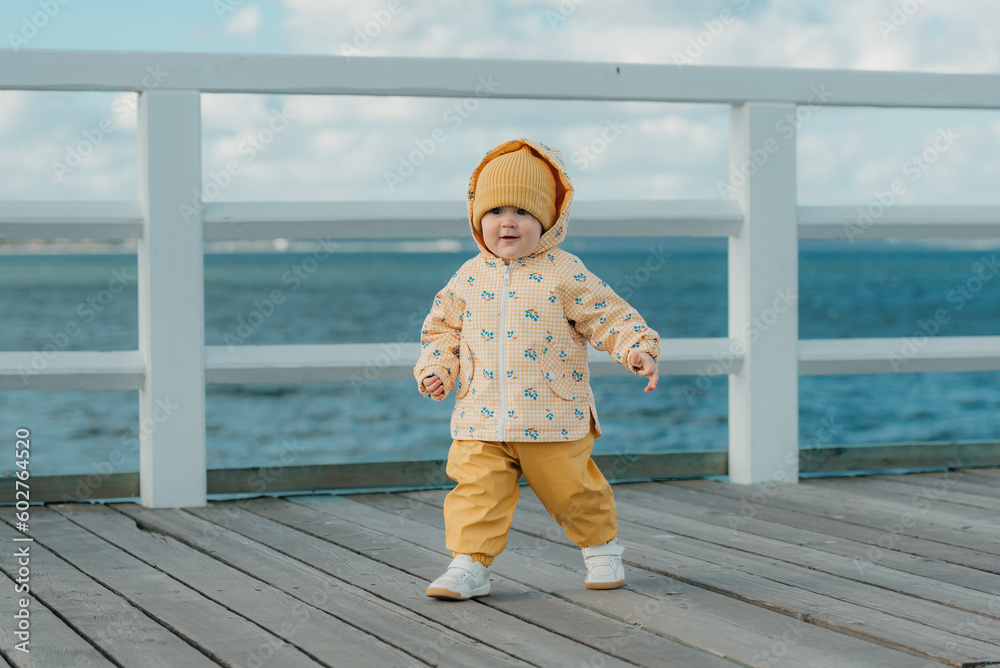 A toddler in a yellow jacket and pants walks on the pier