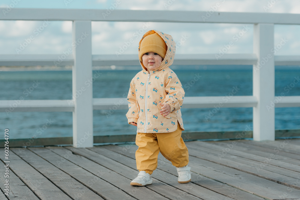A toddler in a yellow jacket and pants walks on the pier