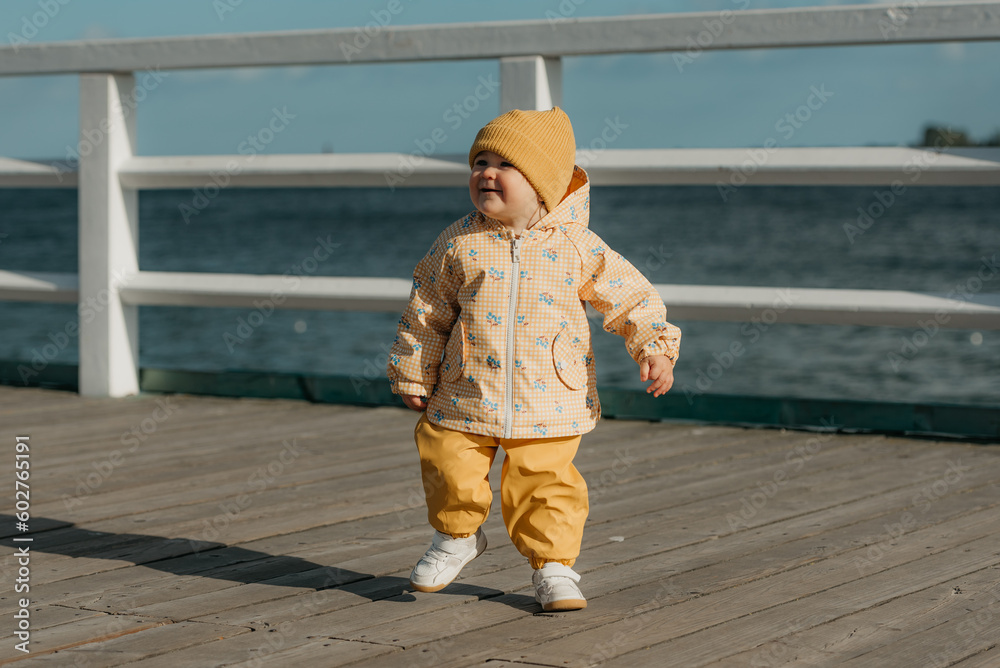 A toddler in a yellow jacket and pants walks on the pier