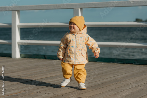 A toddler in a yellow jacket and pants walks on the pier