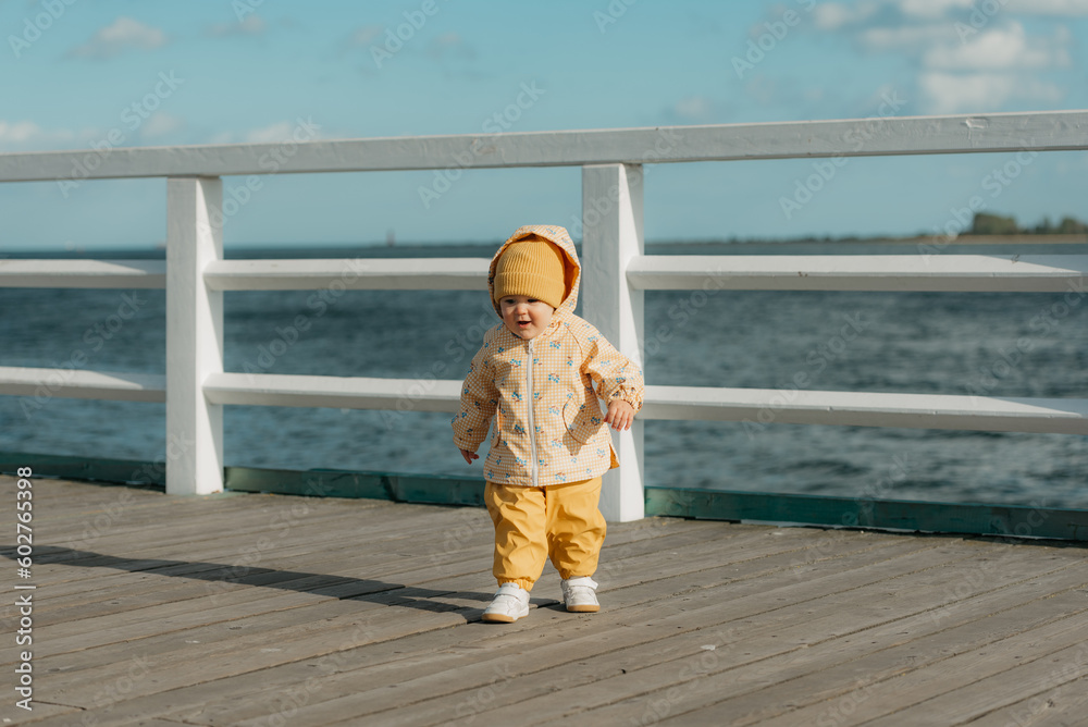 A toddler in a yellow jacket and pants walks on the pier