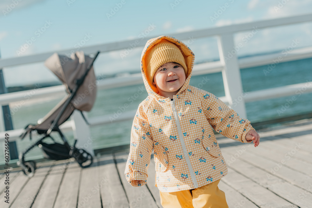 A toddler in a yellow jacket and pants walks on the pier