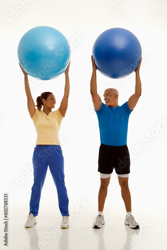 Mid adult multiethnic man and woman holding blue exercise balls over their heads looking at each other smiling.