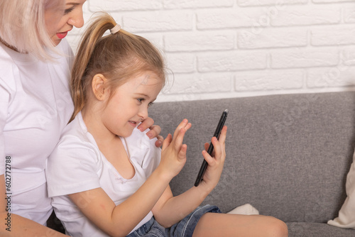 Close up happy mother with little daughter kid using smartphone sitting on couch at home, smiling caring mom and adorable girl child looking at device screen, chatting online, watching cartoons
