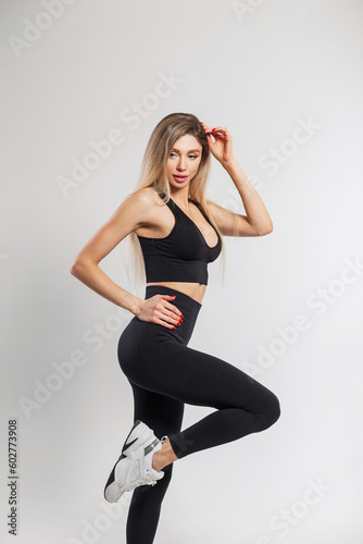 Beautiful athletic fit lady with blond hair in fashionable black sportswear outfit with a top, leggings and sneakers poses on a white background in the studio