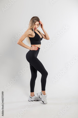 Beautiful sexy fitness model blonde woman with a slim fit body in a fashionable black sportswear with leggings, sneakers and a top on a white background
