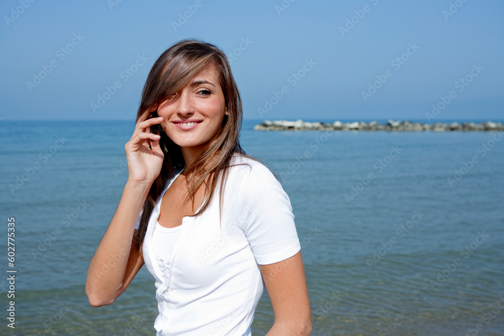 Beautiful sensual woman with long brown hair talking with a mobile phone standing by the ocean in a sunny day Beautiful sensual woman with long brown hair talking with a mobile phone standing by the ocean in a sunny day