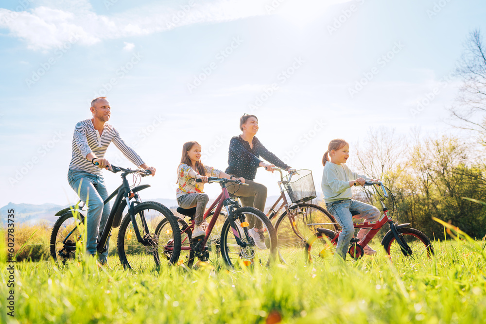 Smiling father and mother with two daughters during summer outdoor bicycle riding. They enjoy togetherness on green high grass meadow. Happy parenthood and childhood or active sport life concept image Smiling father and mother with two daughters during summer outdoor bicycle riding. They enjoy togetherness on green high grass meadow. Happy parenthood and childhood or active sport life concept image