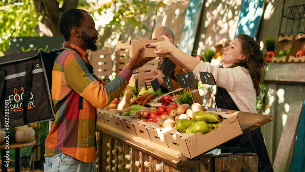 African american deliveryman wearing backpack waiting for order at farmers market counter. Team of farmers delivering fresh organic fruits and vegetables to customers, small business owners.