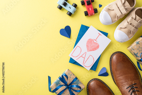 Celebrate Father's Day with a heartwarming top view scene of a little boy congratulating his dad. On yellow backdrop, see dad's leather shoes, baby son's sneakers, toys, handmade postcard and giftbox