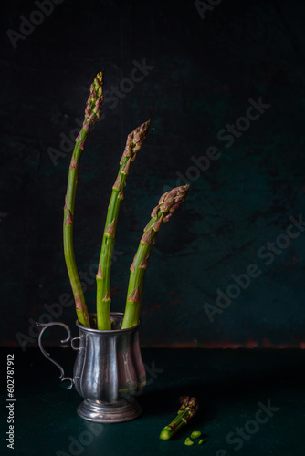 Decorative composition with green fresh asparagus in a cup on a dark green background, book, culinary/food photography, healthy eating concept, vegan/vegetarian diet, still life, blank space for text.