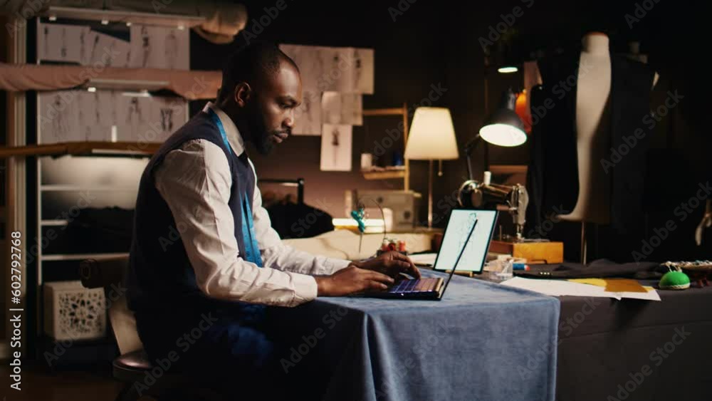 Male couturier using laptop in atelier, searching for clothing items models for inspiration and needlework. Young craftsman and business owner designing new fashion collection. Male couturier using laptop in atelier, searching for clothing items models for inspiration and needlework. Young craftsman and business owner designing new fashion collection.