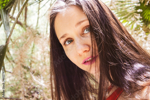 Portrait of a young beautiful enigmatic woman in a playful mood on nature. A girl with cute baby face and blue eyes in tropical garden. Human and nature unity. Straight gaze, look. Good spirits youth.