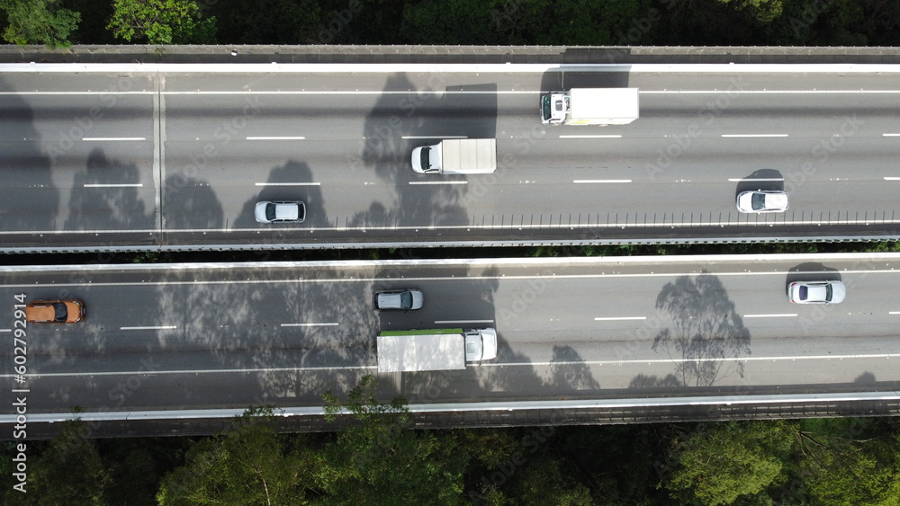 Visão aérea de um carro atravessando a rodovia Mogi Dutra em São Paulo com a sombra das árvores projetando na rodovia. 