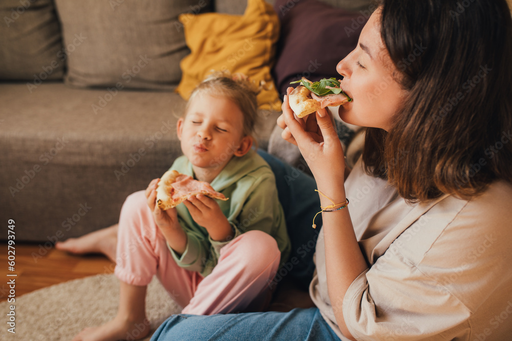Young mother and her little 5 year old daughter eating pizza together in cozy living room. Young mother and her little 5 year old daughter eating pizza together in cozy living room.