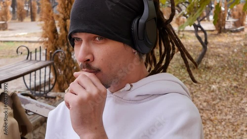 Portrait of an attractive young guy with dreadlocks, an earring in his ear and bracelets on his wrist lighting a homemade cigarette outdoors on a public park bench in autumn and getting satisfied photo