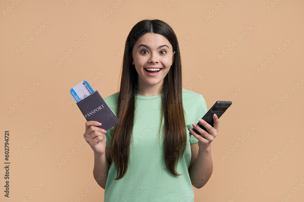 Amazed happy teenager girl looking at camera, posing with mobile phone and flight ticket on beige Amazed happy teenager girl looking at camera, posing with mobile phone and flight ticket on beige