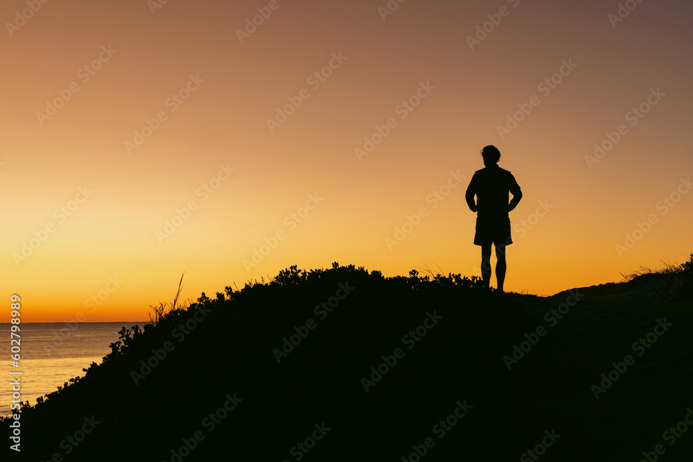 Silhouette of man standing on sand dune overlooking Jimmy's Beach at sunrise. Hawks Nest, NSW Australia