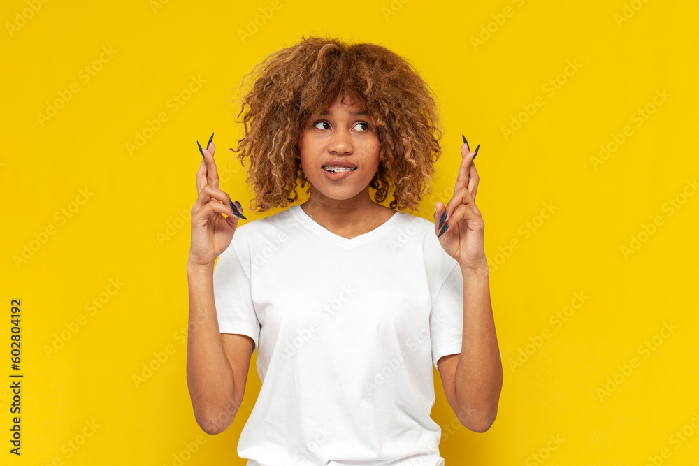 young curly american girl with braces praying and hoping for good luck on yellow isolated background young curly american girl with braces praying and hoping for good luck on yellow isolated background