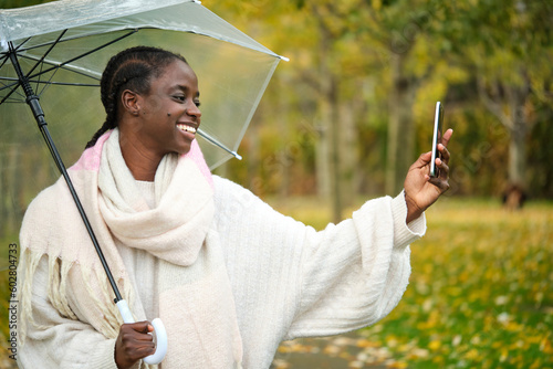 African woman taking a selfie with a smartphone and umbrella smiling in autumn. © Ladanifer