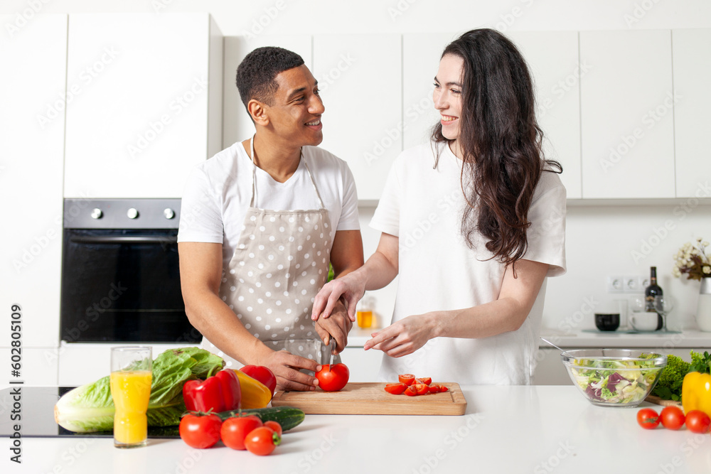 african american man and european woman preparing vegetable and greens salad together in white kitchen