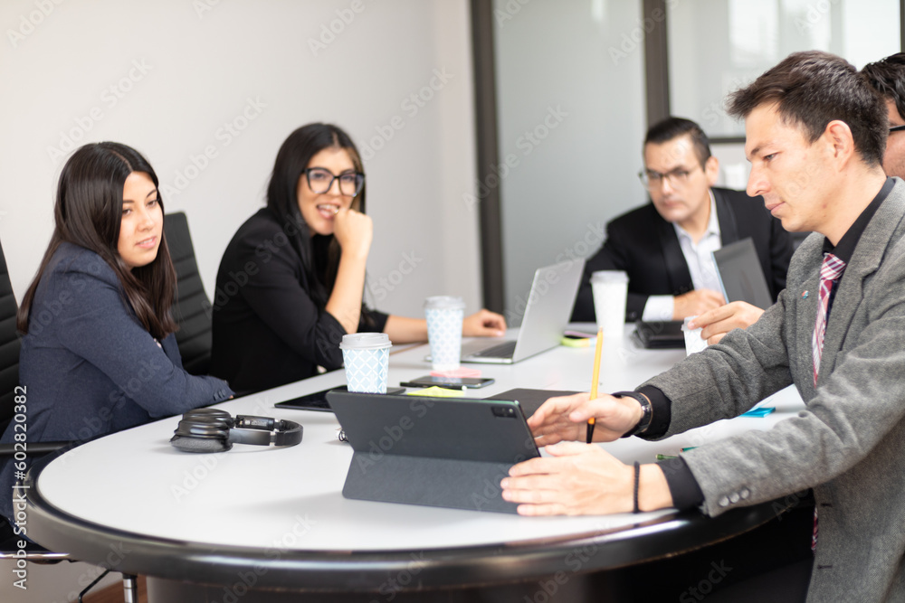 young Caucasian man in a coworking space showing graphics on his tablet to a partner coworkers in a meeting room, busy office ambience, businessmen at work young Caucasian man in a coworking space showing graphics on his tablet to a partner coworkers in a meeting room, busy office ambience, businessmen at work
