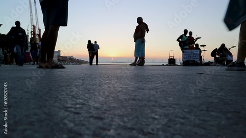 A man sitting down throws out a paper during sunset over Praca Maua, Rio de Janeiro, Brazil. People walk by, a fat man, a couple, a street band plays. photo