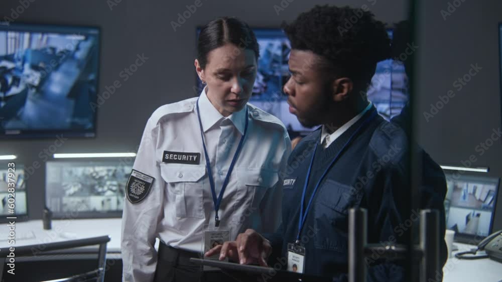 African American male officer uses tablet and talks to female coworker. Multi ethnic employees in security control center. Big digital screens and PC monitors with surveillance CCTV cameras footage.