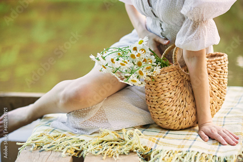 a woman in a light dress sits near the lake and holds a wicker basket with flowers in her hands photo