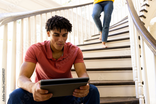 Happy ibracial man sitting on stairs at home using tablet, with legs of woman walking downstairs photo