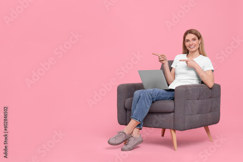 Happy woman with laptop sitting in armchair and pointing at something on pink background. Space for text