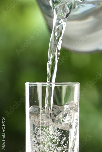 Pouring water from jug into glass on blurred green background, closeup