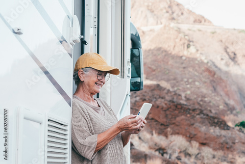 Happy senior woman with eyeglasses standing outside a camper van motor home using mobile phone enjoying travel destination. Smiling elderly lady in alternative vacation transport and tourism concept