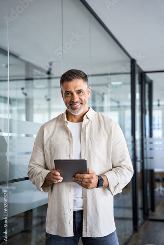 Latin Hispanic mature adult professional business man looking at camera and smiling. Vertical portrait of Indian senior businessman CEO with digital tablet using online application standing in office.
