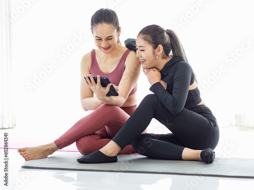Two Asian beautiful healthy fit slim sexy female sporty athlete models friend in sports bra and leggings sitting on yoga mat resting relaxing take break smiling talking looking at tablet together