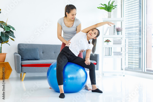 Asian professional fitness trainer training helping pretty healthy fit slim sexy female sporty athlete model in sportwear and leggings using big exercise rubber yoga ball workout training stretching