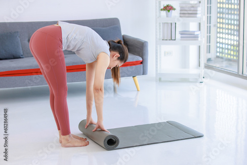 Asian beautiful healthy fit slim sexy female sporty athlete model in sportswear and leggings standing bending head down rolling yoga mat out ready for workout exercising training in living room