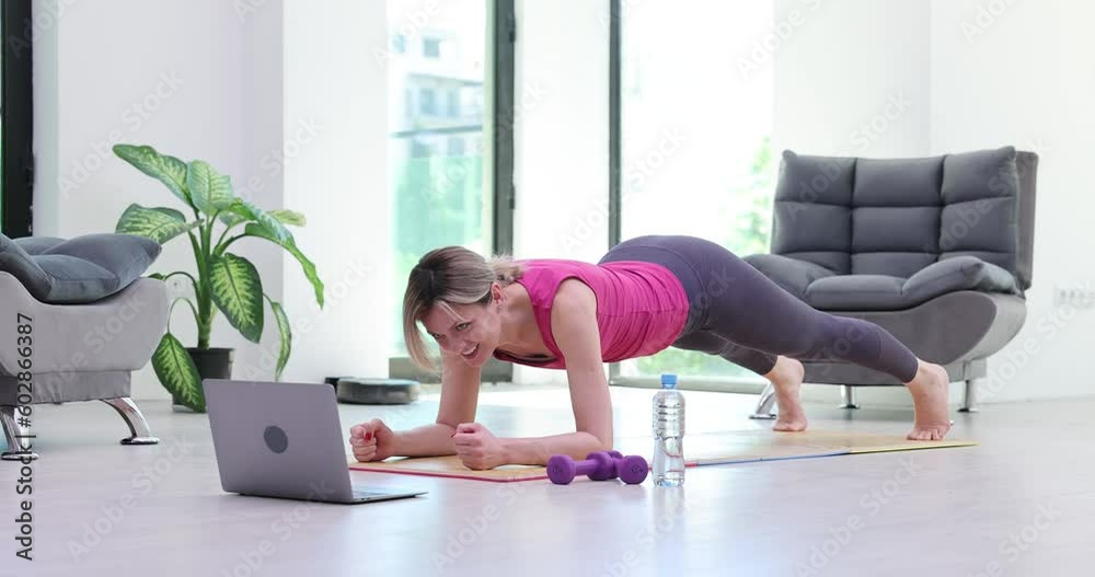Fit woman does yoga plank pose on mat to strengthen body muscles. Female person enjoys online fitness via laptop repeating exercise after coach