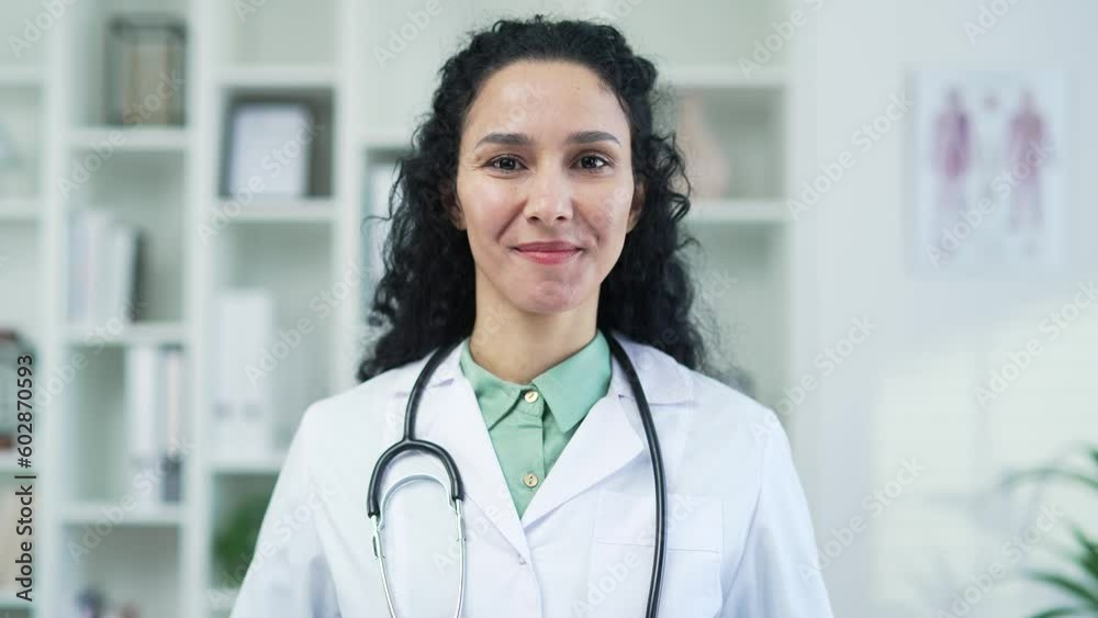 Portrait of smiling female pediatrician in white coat with stethoscope looking at camera in modern hospital clinic. Adult positive brunette woman medical worker doctor poses standing in the office Portrait of smiling female pediatrician in white coat with stethoscope looking at camera in modern hospital clinic. Adult positive brunette woman medical worker doctor poses standing in the office